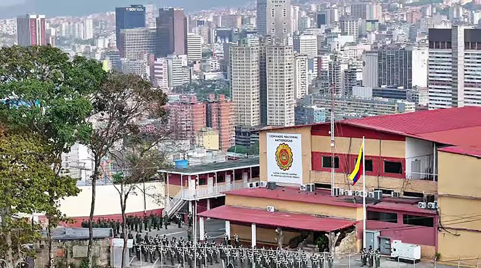 Vista aérea del Comando Nacional Antidrogas de Venezuela, con una bandera venezolana y personal militar en formación, enmarcado por el horizonte urbano de Caracas