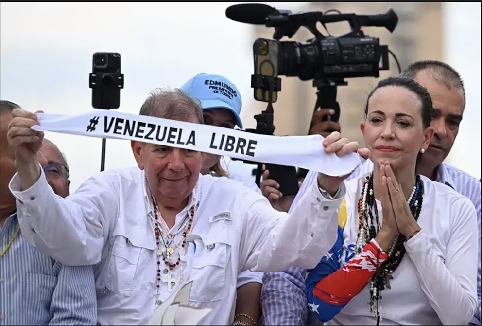 Edmundo González Urrutia y María Corina Machado sostienen una pancarta con el lema '#Venezuela Libre' durante un evento, siendo captados por un equipo de televisión
