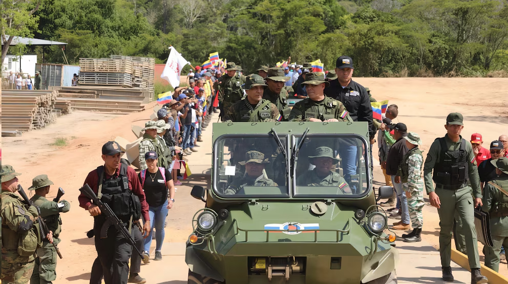 Un vehículo blindado militar avanza por un camino de tierra acompañado por efectivos y una multitud con banderas colombianas.